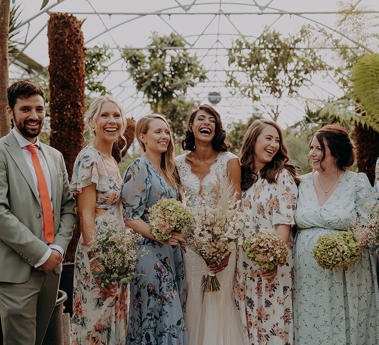 Bride poses with mixed gender bridal party wearing mixed floral dresses and a grey suit