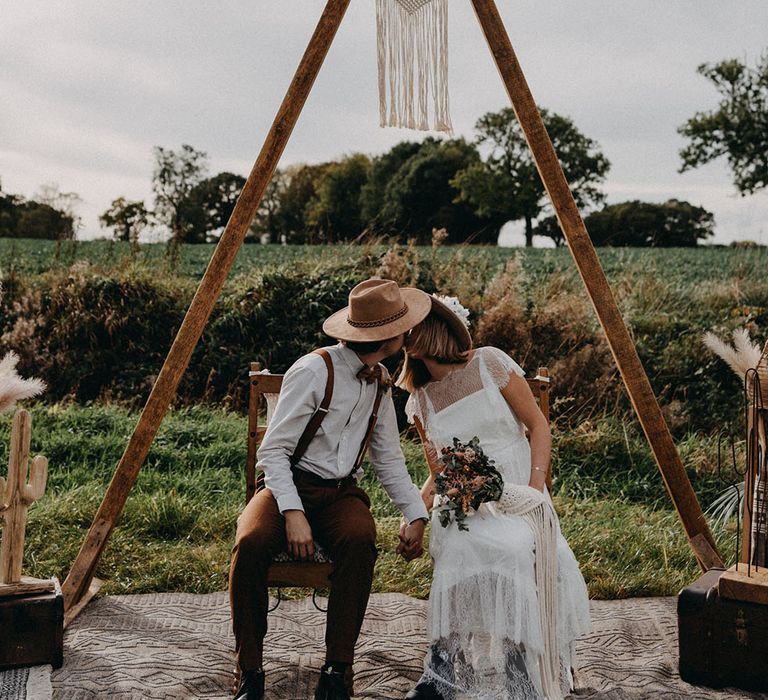 The bride and groom sit together kissing surrounded by handmade decor and dried flowers 