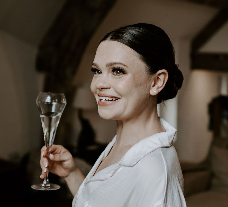 Bride drinks champagne as she gets ready for the wedding in white pyjamas with pink trim and hair in a sleek bun 