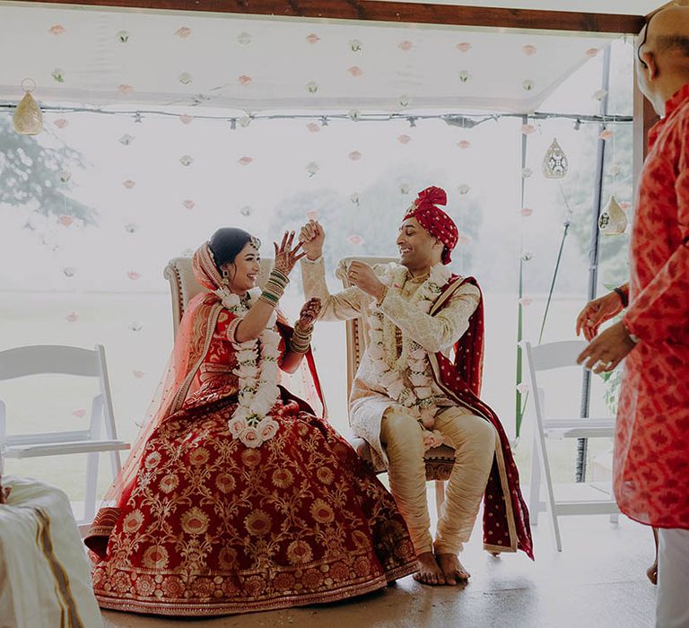 Bride in beautifully embellished red and gold lehenga sits alongside her groom during wedding ceremony 