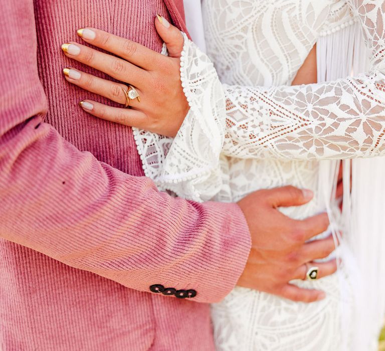 Bride in lace long sleeve wedding dress and diamond wedding ring and groom in pink corduroy suit embracing