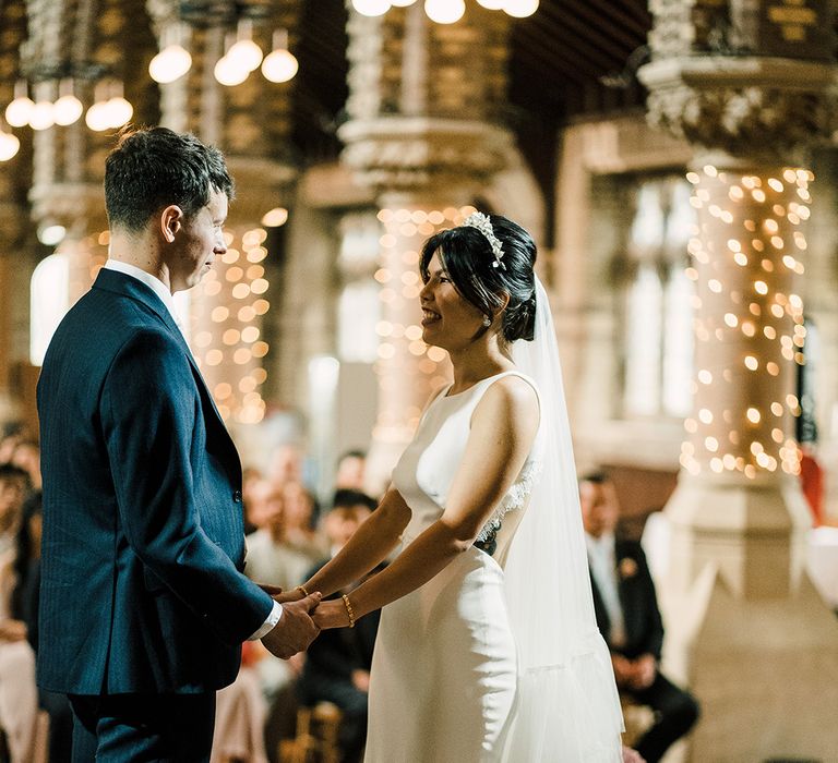 East Asian bride wears embellished bridal crown and fitted open back wedding dress opposite her groom during church wedding 