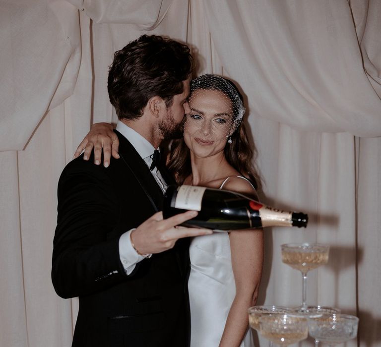 Bride in satin cowl neck wedding dress and birdcage veil and groom in classic black tux pouring champagne into their wedding champagne tower 