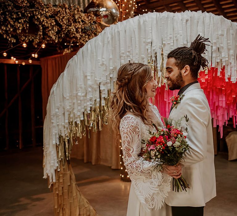 Bride in boho long sleeve lace wedding dress with tassels embracing groom in white blazer with colourful boutonniere, black shirt and black suit trousers standing beside colourful streamer decoration ideas 