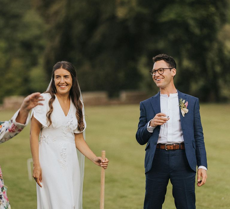 Bride and groom stand sipping champagne playing croquet on the lawn for summer garden party wedding 