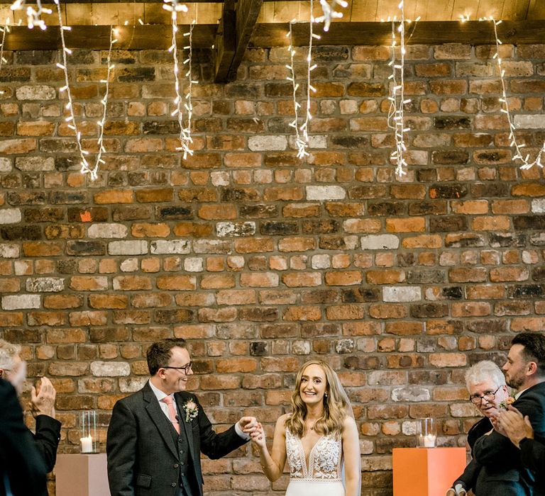 Groom in tartan kilt stands beside his bride in appliqué Mikaella Bridal wedding dress 