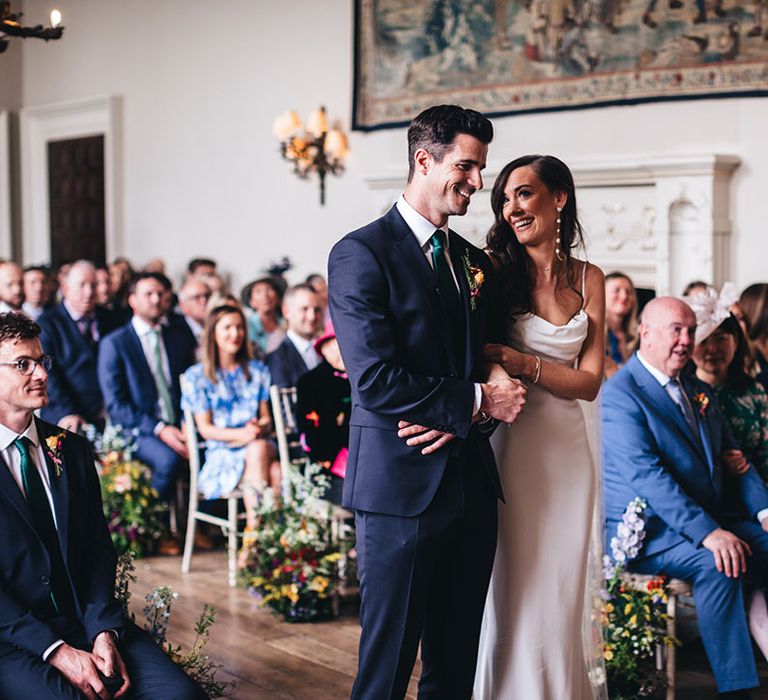 Groom in a green and blue suit stands linking arms with the bride smiling as they participate in their civil wedding ceremony at Elmore Court