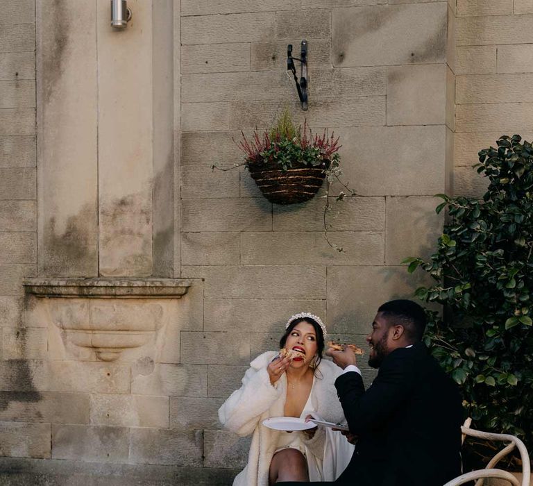 Bride in pearl headband and large faux fur coat eating pizza with groom in black tux at Manor House wedding venue Yorkshire