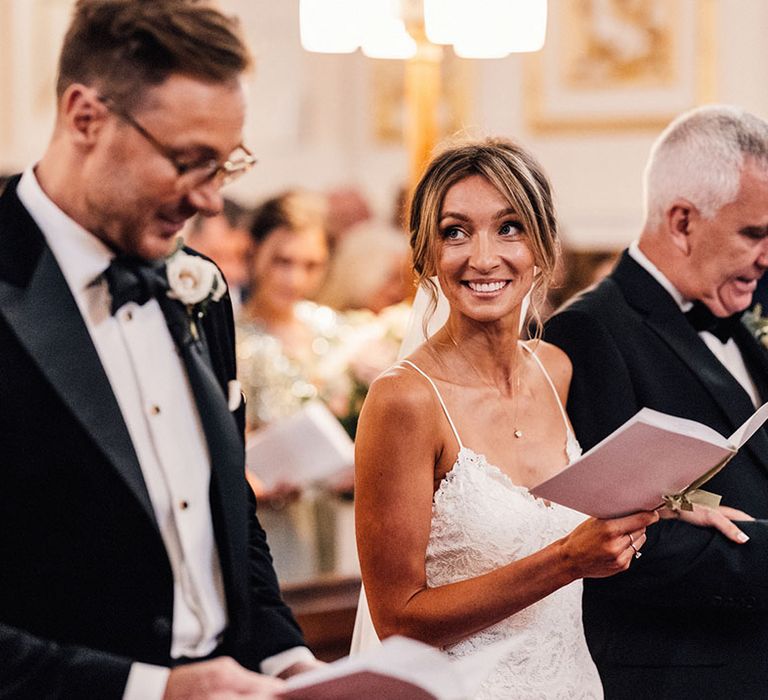 The bride smiles brightly in her fitted lace wedding dress holding a pink order of service wedding booklet at the groom in a black tuxedo