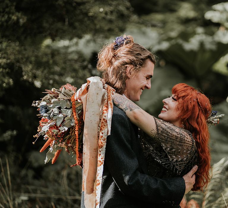 Bride in embellished short sleeve black wedding dress holding orange patterned ribbons embracing groom in dark grey suit 