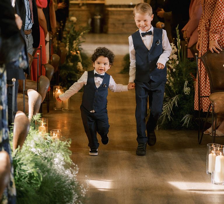 Page boys in navy waistcoat and bow tie walking down the aisle together with pillar candles in hurricane vases and white flowers decorating the aisles 