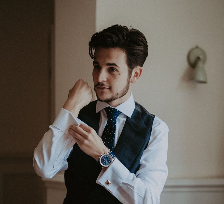 Groom in navy blue waistcoat and tie putting on his cufflinks for the wedding day 