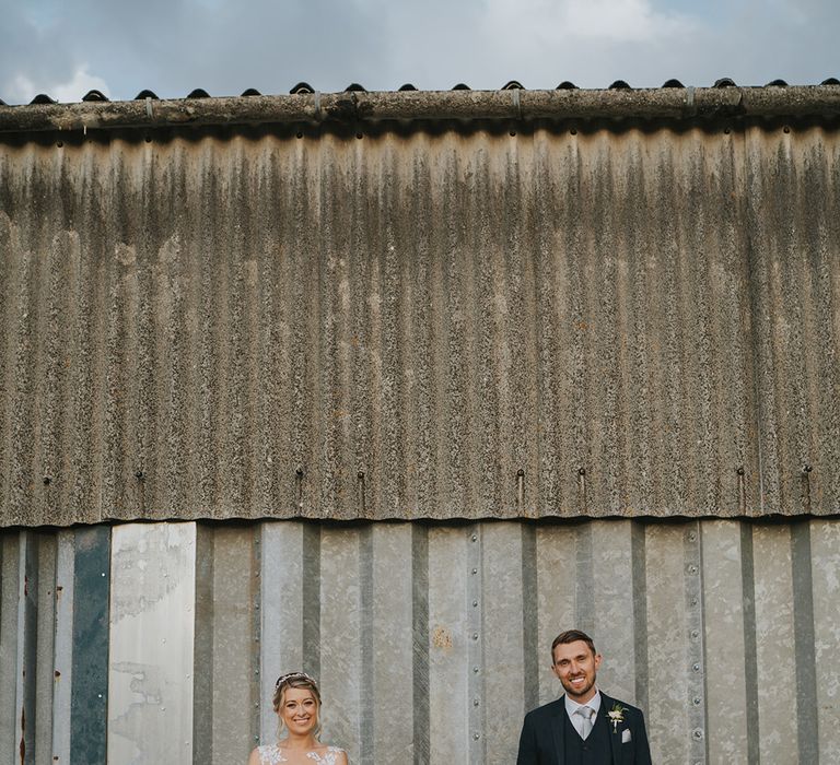 Bride in Dando London wedding dress and groom in dark blue suit stand in front of corrugated building 