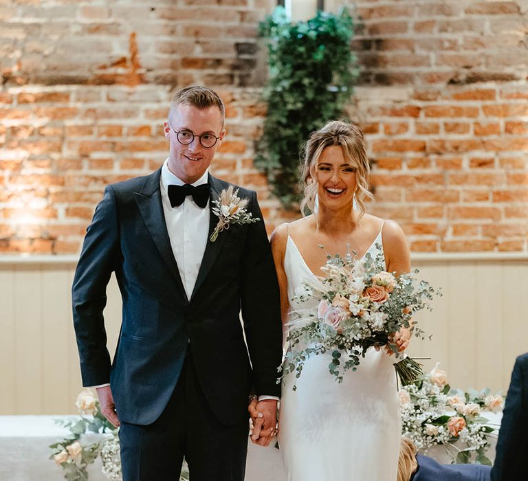 Groom in black tuxedo wearing a dried flower bunny grass buttonhole turns and faces the guests with the bride in a satin slip wedding dress 