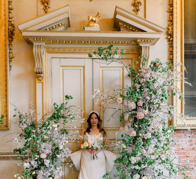 Bride in sleeveless satin wedding dress with large accent bow on the back standing by large rose, baby's-breath, foliage and dried flower floral columns holding classic spring bouquet 