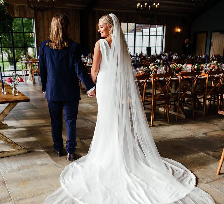 Bride wearing a fitted draped back wedding dress with long train and veil walking around the wedding reception 