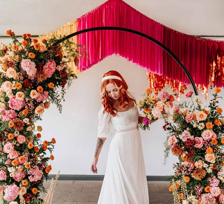 Bride in three quarter length puff sheer sleeve wedding dress with delicate polka dot overlay and square neck standing by circular floral arch with vibrant flowers and foliage attached and pink, red, yellow and metallic wedding streamers behind