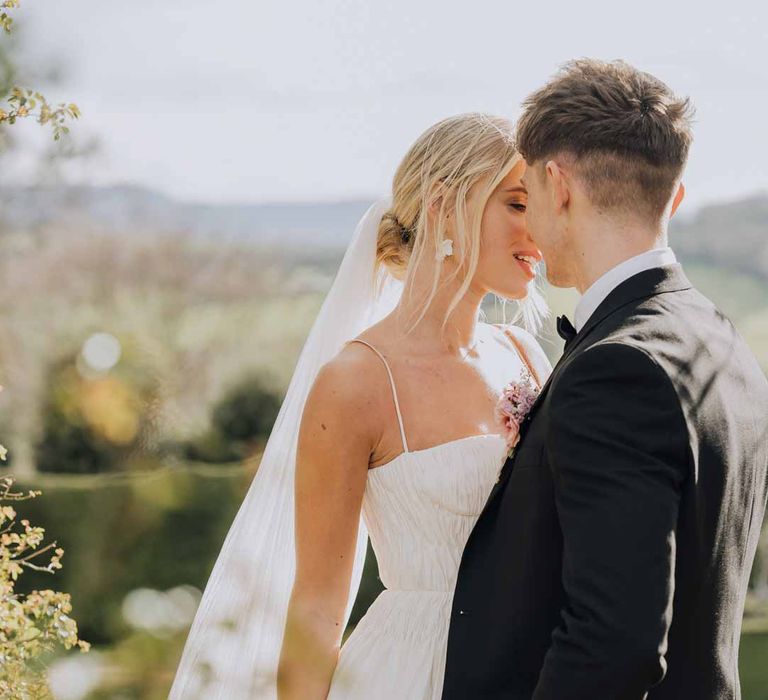 Bride wearing Watters strappy sleeveless princess wedding dress and chapel length wedding veil standing with groom in classic grooms tuxedo and bowtie 