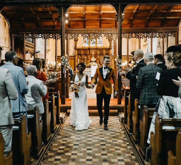 Groom in crushed burnt orange velvet grooms blazer, black bowtie and black suit trousers walking hand in hand with bride in v-neck sleeveless wedding dress with 3D floral applique train holding blush and orange garden roses, poppies and carnations, dried flowers and eucalyptus wedding bouquet walking down the aisle at Brinsop Court wedding