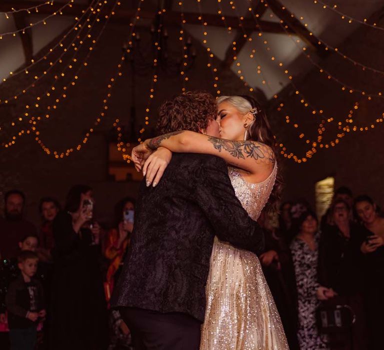 Bride in sleeveless deep v-neck sparkly gold wedding dress and bride in dark velvet suit under fairy lights doing their wedding first dance at Kingscote Barn Tetbury wedding venue