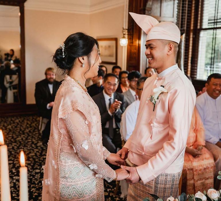 Groom in baby pink htaingmathein holding hands with bride in floral 3D applique pink and rose gold htamane at the alter of Bron Eifion wedding venue 