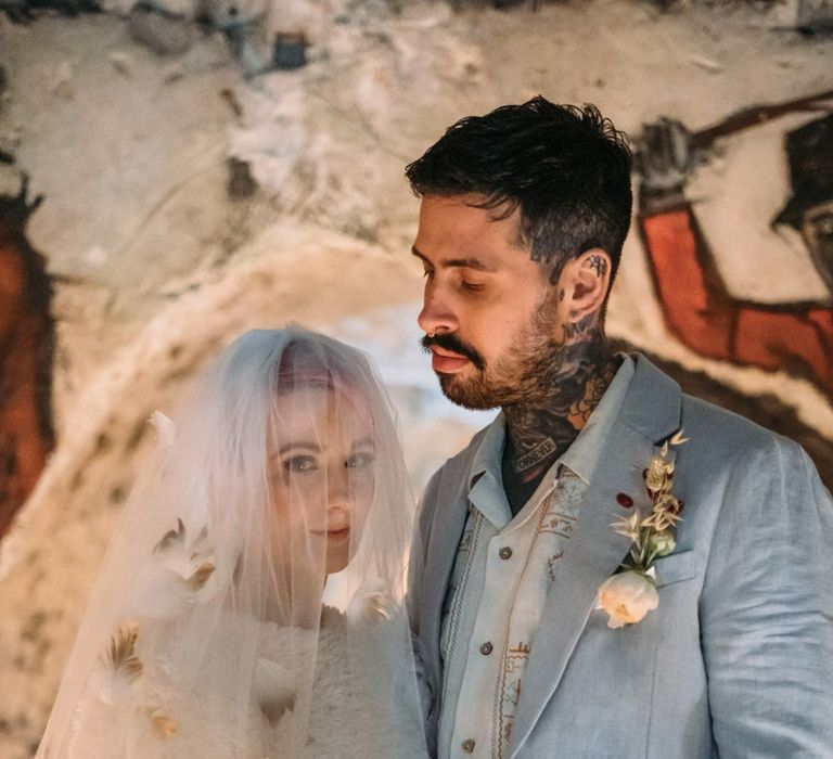 Bride in alternative feather bridal veil standing with groom in baby blue linen suit, personalised embroidered linen shirt and garden rose and pampas grass boutonniere and holding large colourful bridal bouquet 