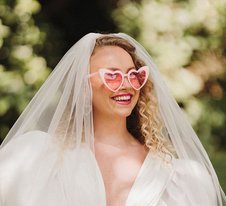 Bride in puff sleeve Watters wedding dress with wedding veil and pastel pink retro wedding sunglasses with pink bouquet 