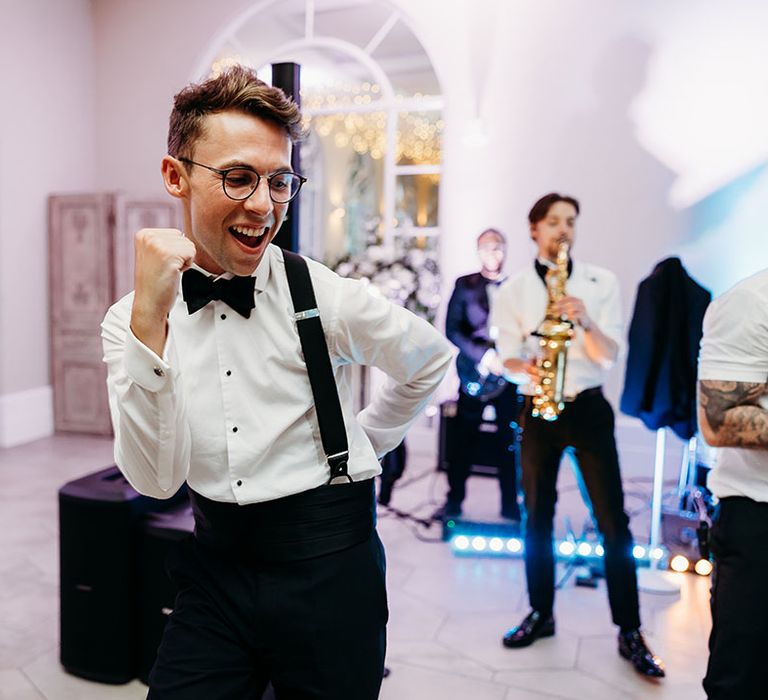 Groom in bow tie and braces dancing to saxophonist from wedding band 