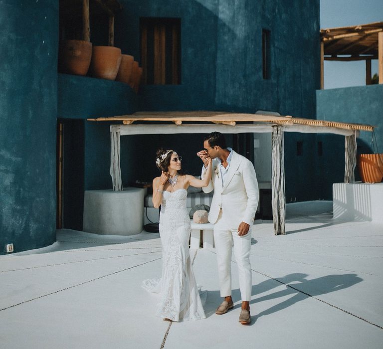 Bride and groom walk in front of blue building at Tigre Del Mar wearing a white boho wedding dress, cream suit and sunglasses