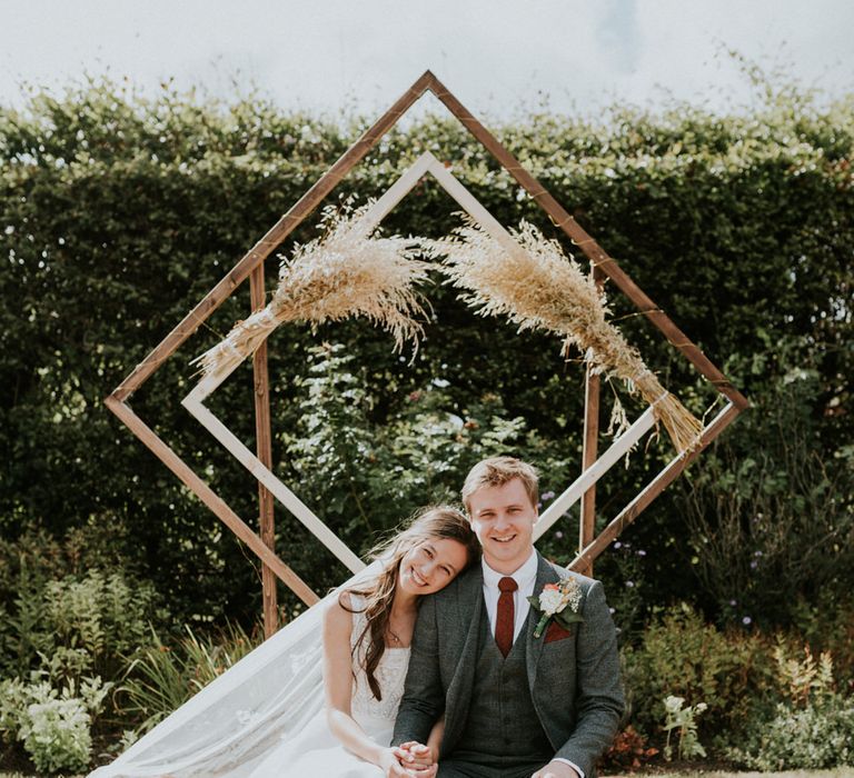 Bride and groom sitting on a hale bale with wooden frame structure behind. 