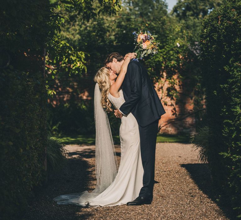 Bride in Justin Alexander wedding dress holding colourful bouquet embraces groom in the grounds of Iscoyd Park