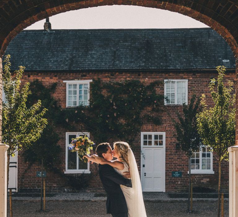 Bride in Justin Alexander wedding dress holding colourful bouquet hugs groom under arch in the grounds at Iscoyd Park