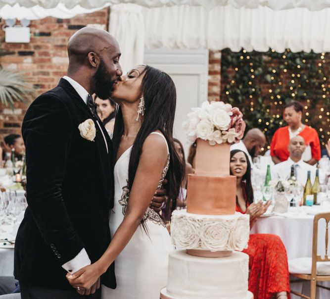 Black bride and groom kissing next to their white and rose gold wedding cake surrounded by individual cupcakes 