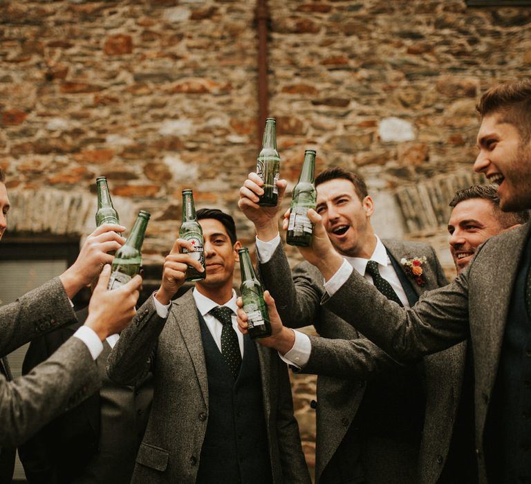 Groomsmen in grey wool suits with navy waistcoats and polka dot ties drinking bottled beer on the wedding morning 