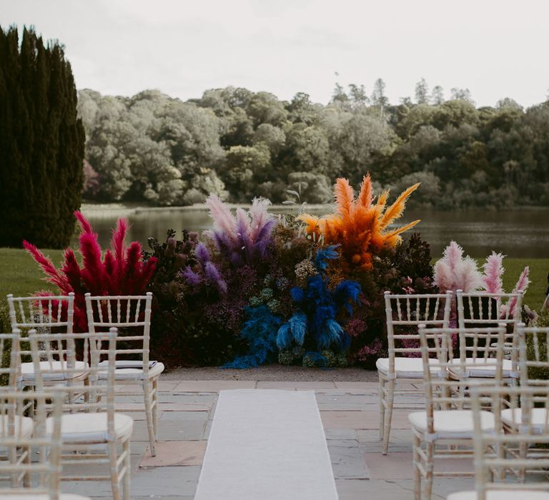 Vibrant multi coloured pampas grass floral installation at the end of the lakeside aisle with white bamboo wedding chairs