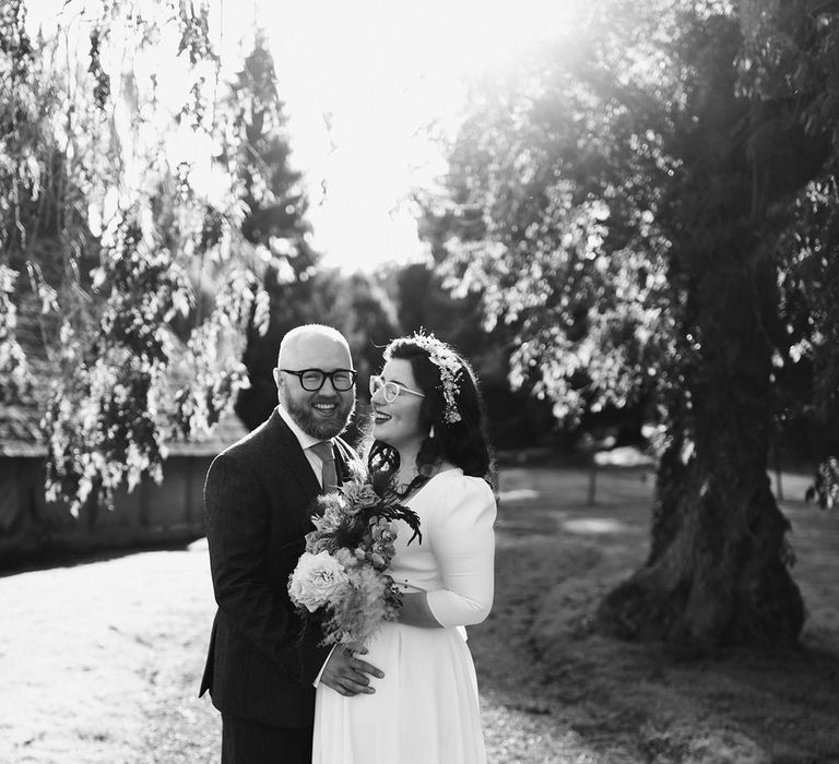 Groom in navy suit and yellow tie stands with bride in white cat eye glasses and bridal headband holding white and pink rose and pampas grass bouquet