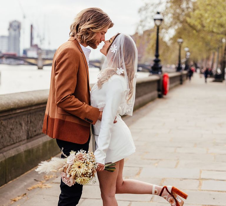 Groom in a rust coloured blazer embracing his bride by the Thames in a short blazer dress with appliqué veil, platforms and retro socks 