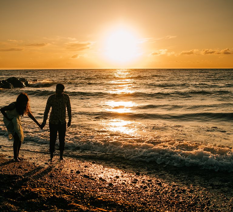 Sun sets across the sea as bride & groom walk along the beach