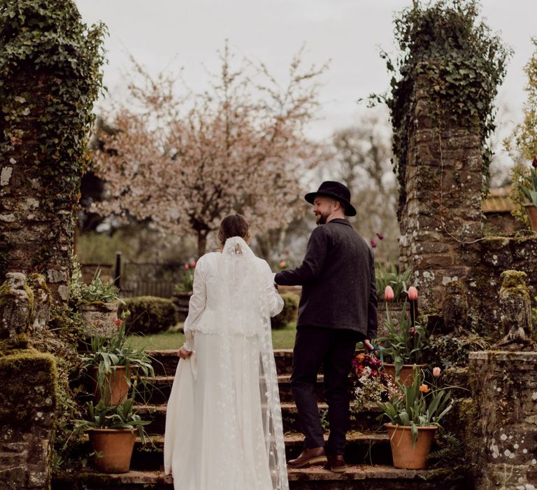 Bride in white Charlie Brear wedding dress with slit and chapel length daisy applique veil walks arm-in-arm with groom in grey tweed suit up garden steps at garden party wedding in Devon