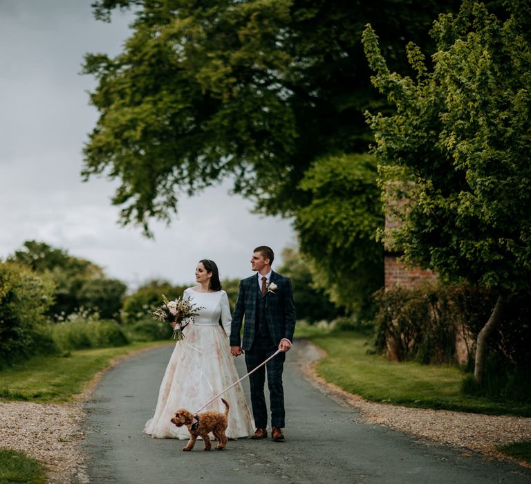 Rural bride and groom portrait with the bride in a floral skirt wedding dress and the groom in a navy check suit walking their pet cockapoo 