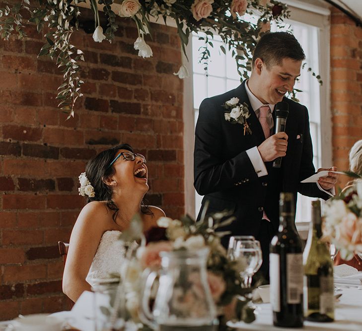 A bride in glasses laughs as her husband reads his wedding speech.