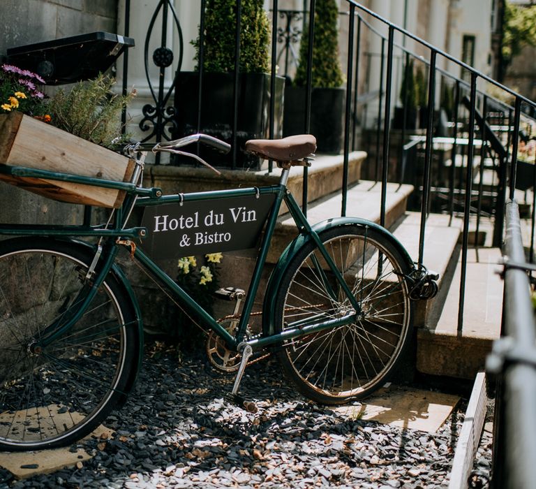 Dark green bicycle with Hotel du Vin & Bistro sign and wooden box filled with flowers attached on flint stones in front of the Hotel du Vin Harrogate