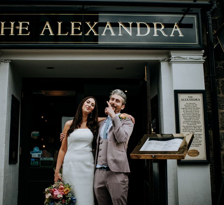 Bride in white strapless Rebecca Vallance dress holds multicoloured bridal bouquet and has arm around groom in brown Moss Bros suit and blue tie as he points to pub sign reading 'The Alexandra' in Harrogate 