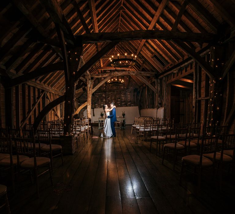 Bride & groom stand in large barn whilst lights twinkle around them