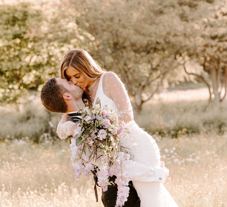 Groom lifts bride as they stand within fields during golden hour on their wedding day