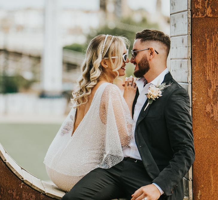 Bride in a sparkly wedding dress, pearl headband and white sunglasses leaning in to kiss her husband in a black suit, white shirt and aviator glasses 