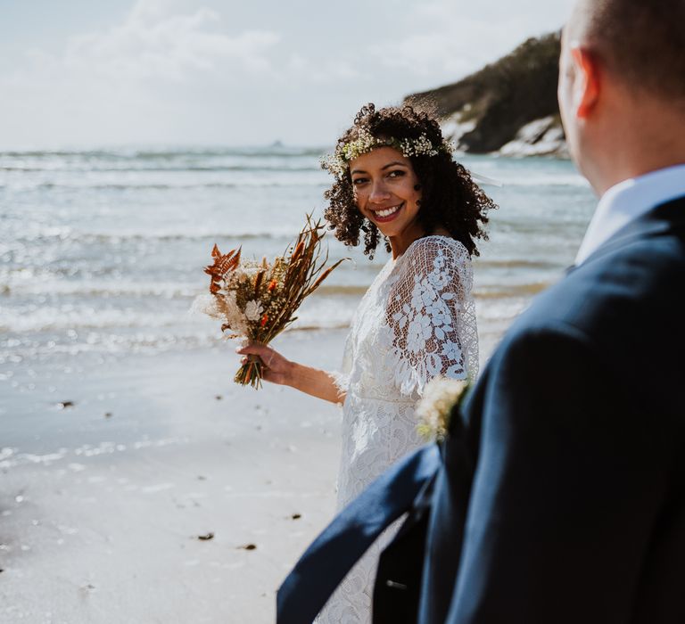 Bride walks across the beach as she looks back at her groom whilst holding dried floral bouquet