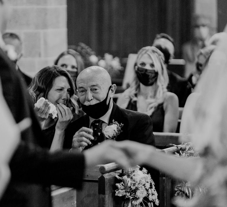 Two wedding guests with face masks on get emotional as bride and groom hold hands at altar during church ceremony