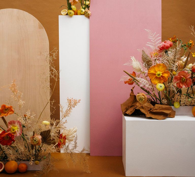 Wedding flowers on plinths including pink and yellow poppies, roses, dried palms, dried ferns and citrus fruit