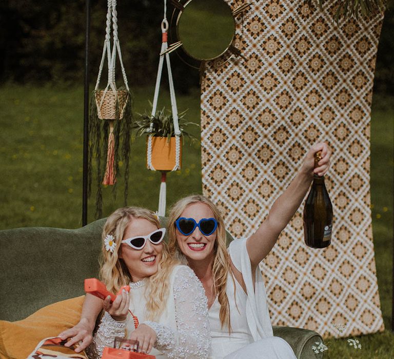 Two brides sitting on a love seat with retro print fabric backdrop, macrame hanging plant pots, and wildflowers